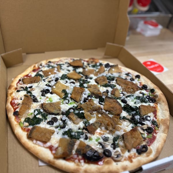 A Jonuzi's Pizza with white sauce, spinach, olives, onions, and fried eggplant in a cardboard box.