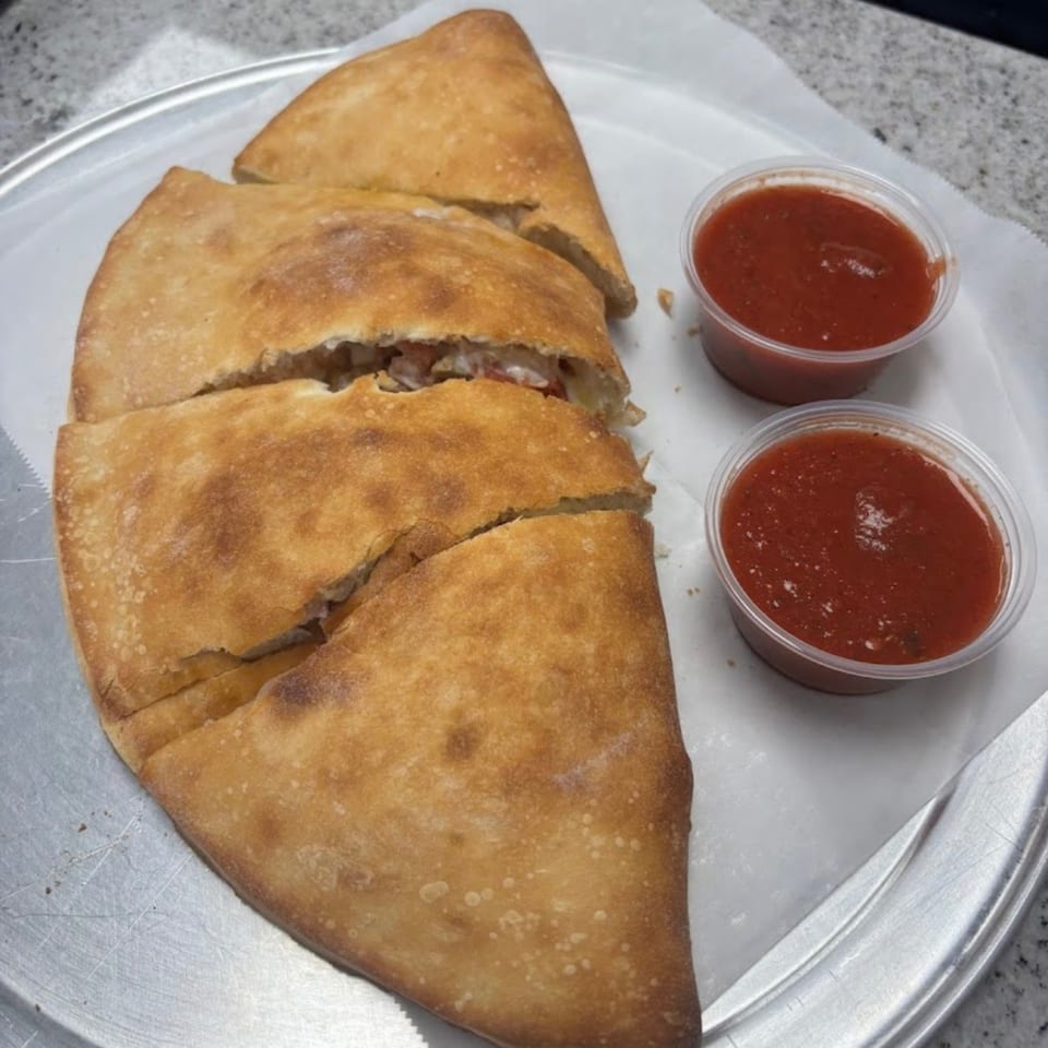 A sliced Jonuzi’s Pizza calzone with two containers of red sauce on a metal tray.